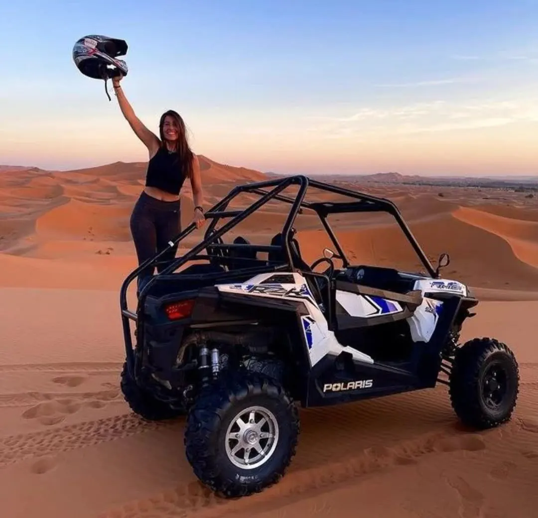 Viaggiatrice in piedi su un buggy, con il casco in mano, in cima a una duna al tramonto