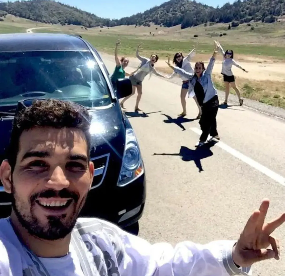Conductor junto a la furgoneta del tour privado, con cinco viajeras saludando en una carretera de montaña de Marruecos