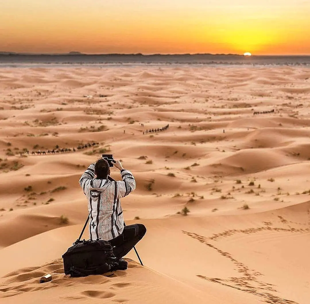 Fotografo con treppiede in cima a una duna, con carovane di cammelli in lontananza e il sole all’orizzonte