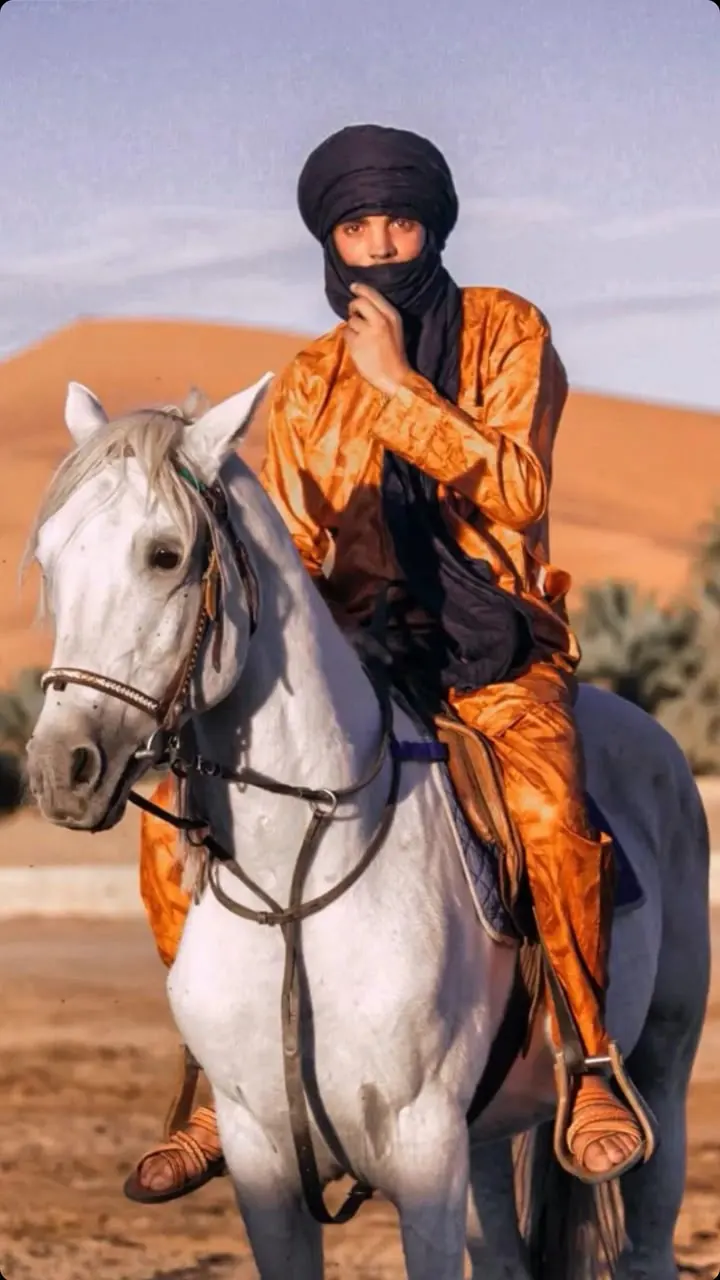 Cavaliere con turbante e tunica arancione su un cavallo bianco, con le dune di Merzouga alle spalle